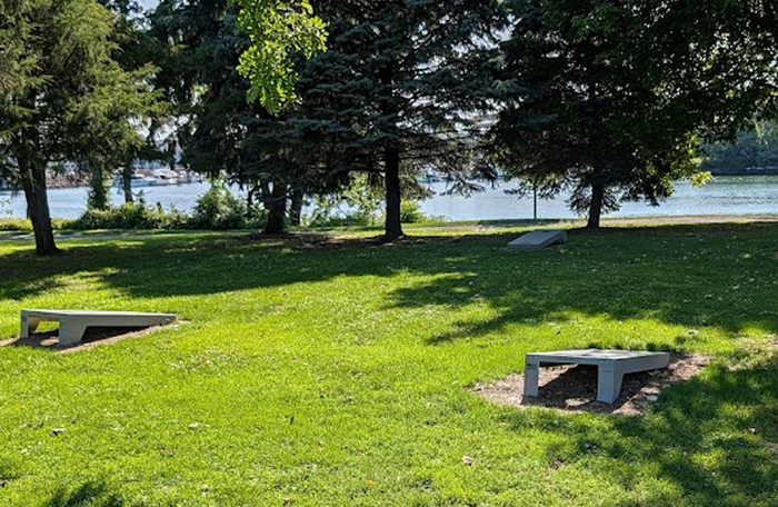 Permanent cornhole boards installed on a grassy lakeside park beneath mature trees.