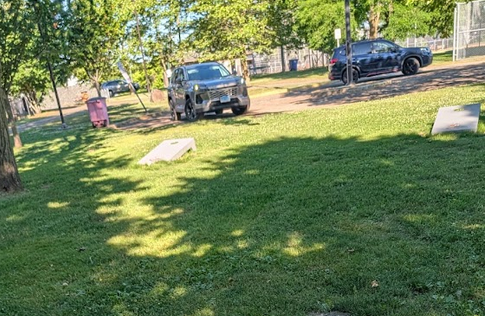 Cornhole boards set up on a shaded lawn near parked vehicles and a neighborhood street.
