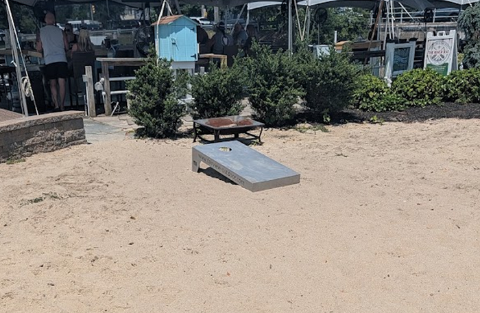 Gray cornhole board positioned on sand beside an outdoor dining and gathering area.